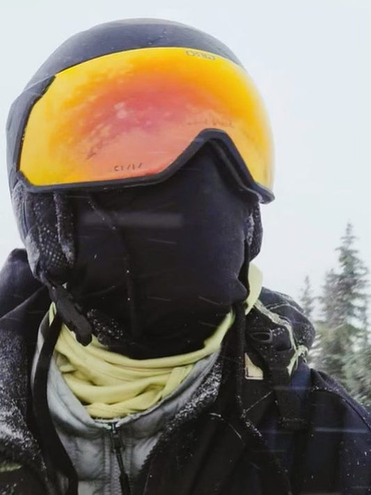 Man seen in ski gear ,with black face covering and holographic orange and red goggles with yellow undershirt followed by grey sweater 